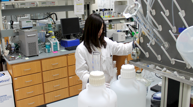 A woman works at a lab bench, pipetting liquid into a tall flask